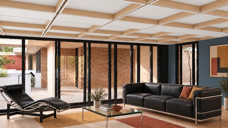 Ein Wohnraum unter der Kassettendecke aus Holz: links eine Liege aus Stahlrohr und Leder, rechts ein schwarzes Ledersofa vor blauer Wand, dazwischen ein Glastisch auf einem Teppich; hinter der schwarz gerahmten Glaswand ein Hof mit Wänden aus Backstein und einem Holzdeck.