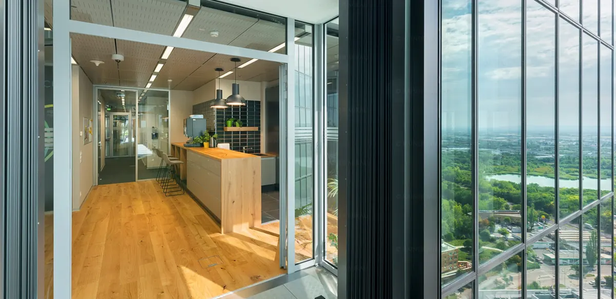 Offene Bürofläche mit Teeküche und Theke aus Holz, dahinter raumhohe Verglasung mit Blick über die Stadt